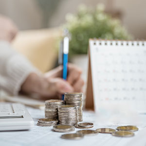 Stacks of coins and a calendar on a desk. A person writes in the background.