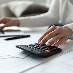 Hands using a calculator on a desk with a laptop and documents.