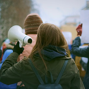 Person in green jacket holding a megaphone at outdoor protest, surrounded by demonstrators with signs in cold weather