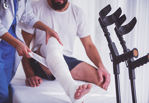 Doctor wrapping cast on injured leg of man with crutches nearby